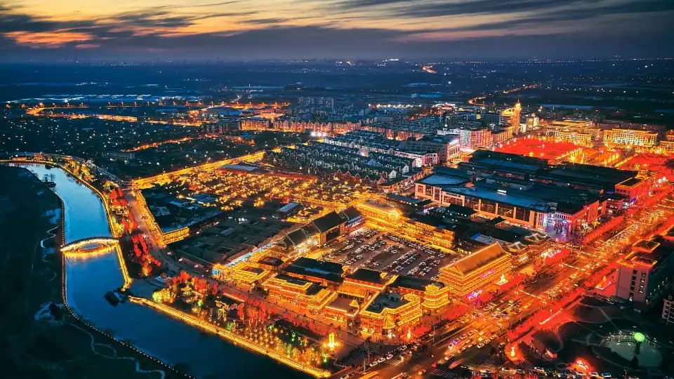 Aerial view of the canal and illuminated Yangliuqing streets at dusk, with modern buildings beyond the old quarter.